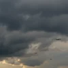 A dramatic sky over the Voronezh region with dark, ominous clouds and faint silhouettes of unmanned aerial vehicles (drones) visible in the distance, conveying a sense of aerial threat and tension, realistic atmospheric lighting, no text or symbols