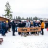 A bustling outdoor labor market in a remote Siberian colony near Arkhangelsk, with diverse groups of people in work attire (mechanics, builders, farmers) interacting, negotiating, and exchanging tools or documents. Include a wooden sign reading 'Биржа труда' (Labor Exchange) in Cyrillic, surrounded by snow-covered ground, wooden huts, and distant pine trees under a cold, overcast sky.