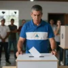A dramatic scene of a Nicaraguan president standing before a closed ballot box with a stern expression, surrounded by empty voting booths and a crowd of citizens looking on with concern. The setting is a dimly lit polling station with broken windows and barbed wire visible in the background. The atmosphere conveys tension and the end of democratic processes.