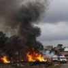 A dramatic scene of a forest fire in a Spanish countryside, with towering flames and thick black smoke rising over pine trees, a distant silhouette of a small village, emergency vehicles with flashing lights approaching the scene, and a somber, cloudy sky, conveying the seriousness of the tragedy without showing graphic details or victims.