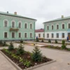 A picturesque courtyard in Rybinsk undergoing renovation. Focus on the architectural details of the buildings, the landscaping work in progress (new plants, paving), and the overall atmosphere of revitalization. Include workers in the background, but keep them small and not the main focus. Show a mix of old and new elements harmoniously blended.