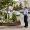 A city official standing on a street corner in Yaroslavl, Russia, explaining the importance of mobile landscaping to residents. The scene includes greenery such as trees, shrubs, and planters along the sidewalk, with people listening attentively. The setting is urban with buildings in the background, and the atmosphere is bright and inviting.