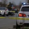 A somber, slightly blurred image depicting a police car at the scene of a traffic accident in Perm, Russia. The scene should include emergency lights flashing, police tape cordoning off the area, and a general sense of sadness and loss. Focus on conveying the gravity of the situation without depicting any graphic details. The background should show a typical urban street scene with buildings and possibly some trees.