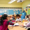 A modern elementary school classroom with children engaged in interactive learning activities, such as group discussions and hands-on experiments, without any traditional homework materials like textbooks or worksheets. The classroom is bright and colorful, with digital screens and educational games visible. The children appear happy and focused on collaborative learning.
