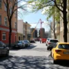 A wide shot of a street in the Krasnooktyabrsky district of Volgograd, Russia, on a sunny day. Several cars are parked along the street. A construction site is visible in the background with workers in safety vests. The scene depicts a temporary suspension of activity, with construction paused.