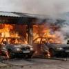 A scene of a fire in a garage in the Roslavl district, with two burning cars inside. The garage is engulfed in flames, and smoke is billowing out. Firefighters are present, actively working to extinguish the fire. The surrounding area shows signs of damage and urgency.