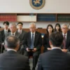 A solemn and respectful scene depicting a group of people gathered for a memorial service in a university or academic setting. The group includes professors, students, and colleagues, all dressed in formal attire. The setting is a hall or auditorium with academic decorations, such as books, diplomas, and university symbols. The atmosphere is one of quiet reflection and mourning.
