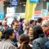 A bustling job fair scene in Chelyabinsk, Russia, with diverse groups of people networking and interacting. In the foreground, a group of family members (multi-generational) are engaged in conversation with a smiling employer representative from a local company. The background shows various booths with company banners, some displaying Russian text, and other attendees. The atmosphere is professional yet warm, with natural daylight illuminating the outdoor or large indoor venue. Focus on the inte