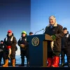 A dignified portrait of Russian President Vladimir Putin standing at a podium, smiling warmly while congratulating workers of the fishing industry on their professional holiday. Behind him, a subtle backdrop shows a calm sea with fishing boats on the horizon and stylized waves. Workers in traditional fishing attire — rubber boots, oilskins, and gloves — stand respectfully in the foreground, some holding fish or nets. The atmosphere is solemn yet celebratory, with soft golden-hour lighting emphas
