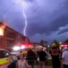 A residential building in a regional city with visible fire and smoke coming from a window after a lightning strike. Emergency services, including firefighters and ambulances, are arriving at the scene. People are evacuating the building, some looking shocked or concerned. The sky is dark with storm clouds, and there is a visible lightning bolt striking the building.