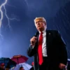 Donald Trump standing confidently outdoors during a thunderstorm, lightning flashing in the dark sky, rain falling, wearing a dark suit and red tie, microphone in hand, addressing a crowd of Americans visible in the background under umbrellas, dramatic lighting, resolute expression, American flag subtly visible in the distance