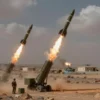 A dramatic, realistic scene showing Iranian military forces launching missiles toward distant American military installations in a Middle Eastern desert landscape. In the foreground, Iranian missile launchers are positioned on a rocky outcrop, with plumes of smoke and fire as rockets ascend. In the background, faint silhouettes of U.S. bases with radar dishes and armored vehicles are visible on the horizon, under a dramatic sky with clouds of dust and distant explosions, conveying tension withou