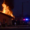 A nighttime scene showing a private house engulfed in flames in the city of Verkhnyaya Salda, with visible fire and smoke, emergency vehicles with flashing lights parked nearby, and people evacuating or watching from a safe distance, all rendered in a realistic photographic style