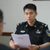 A serious and professional scene depicting a law enforcement officer in uniform, possibly in an office or interrogation room, with a neutral background. The officer is holding a file or document related to the case, with a focused and determined expression. The setting should convey the gravity of the situation, with subdued lighting and a formal atmosphere.