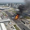Aerial view of a modern city with several transportation hubs, including airports, ports, and train stations, under attack with visible smoke and explosions. The scene should depict the chaos and destruction caused by the strikes, with emergency vehicles and people evacuating the area.
