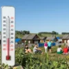 A sunny day in a rural area of Voronezh Oblast, Russia, with a thermometer showing 32 degrees Celsius. The scene includes a small village with traditional wooden houses, lush green fields, and a clear blue sky. People are seen enjoying the hot weather, some sitting under the shade of trees, others walking around in summer clothing.