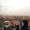 A wide shot of a residential area in Yaroslavl, Russia, with several houses visible. The sky is overcast and a hazy, yellowish-brown smog hangs in the air, suggesting pollution. People are shown wearing masks. Focus on the atmosphere of pollution and its impact on the city.