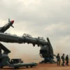 A futuristic military scene with advanced anti-aircraft systems and outdated missiles being loaded onto a launcher. Soldiers in tactical gear operate the equipment under a cloudy sky, with smoke and explosions in the background. The focus is on the contrast between modern technology and obsolete weaponry.