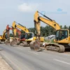 A wide shot of a road construction site in Chelyabinsk, Russia. Heavy machinery like excavators and bulldozers are working on a road. Workers in safety vests are visible. The scene should depict ongoing road repair and improvement.