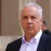 A serious-looking man in a suit standing outside a courthouse. He has a stern expression. The background shows the courthouse building and a few blurred figures of people. Focus on the man's face and upper body.