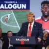 A high-quality image depicting a press conference or a sports event where Donald Trump is speaking about the red card given to Balogun during a championship match. The scene should include a podium with Trump speaking into a microphone, a large screen or banner displaying Balogun's name and a red card, and a crowd of spectators or journalists in the background. The atmosphere should be serious and focused on the topic of sports and politics.