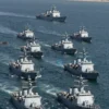 A dramatic maritime scene showing twelve naval ships in the process of being redirected by military forces, with clear visual indication of a blockade enforcement operation in the Strait of Hormuz, featuring Iranian coastal geography and naval vessels in action