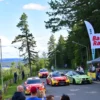 A vibrant outdoor scene in Viipuri featuring a rally event with colorful cars racing on a scenic road surrounded by trees and spectators watching from the sides. Include banners with 'Baltic Rally' text, musical elements like speakers or a band in the background, and a lively atmosphere with people enjoying the event.