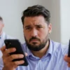 A serious adult man in a modern medical office looking concerned while holding a smartphone displaying a downward trending graph, with a doctor in the background reviewing medical charts, soft clinical lighting, realistic details, no text or numbers visible