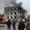 A solemn scene depicting the aftermath of a rocket strike in a residential area of Belgorod region. Show a destroyed building with debris scattered around, emergency services working at the site, and a group of people mourning the loss of a civilian. The image should convey a sense of tragedy and resilience.