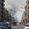 A wide shot of a city street in Iran, showing smoke rising from multiple locations. Focus on the urban environment, buildings, and possibly some emergency vehicles in the distance. The overall mood should be somber and reflective of the event. Avoid depicting any specific damage or casualties.