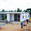 A modern modular medical facility being constructed in a rural village setting, with construction workers and equipment visible. The facility should have a clean, contemporary design with large windows and a welcoming entrance. The village in the background should have traditional houses and a serene landscape with trees and fields.