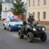 A dynamic street scene in Ryazan, Russia, showing a police car with flashing lights in pursuit of a quad bike (ATV) speeding down a paved road. The quad bike rider is wearing casual clothing and a helmet, leaning into a turn. Buildings with Russian architectural style line the street, with trees and street signs visible. Motion blur emphasizes speed, and the atmosphere is tense but not violent. Daytime setting with clear visibility.