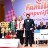 A joyful family from Ulyanovsk participating in a competition, dressed in colorful and festive clothing, standing on a stage with a large banner that reads 'Family Competition' in the background. The family members are smiling and holding hands, with a trophy and a large check for 5 million rubles visible on a table nearby. The scene is bright and cheerful, with a crowd of spectators in the background, clapping and cheering.