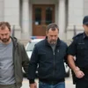 A group of three individuals being escorted by law enforcement officers in a public setting, with a focus on the serious and somber atmosphere. The scene should include elements that suggest a legal or judicial context, such as a courthouse or police station in the background. The individuals should be depicted in a way that emphasizes their current situation, with neutral expressions and appropriate attire.