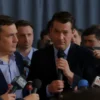 A tense press conference scene with journalists holding microphones, a stern-looking official being questioned, subtle signs of discomfort in the crowd, muted colors, realistic lighting, no text or logos