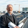 A professional portrait of a middle-aged Russian man, Vasiliy Anokhin, standing confidently in front of a modern urban backdrop showing ongoing communal infrastructure projects: newly built water pipelines, electric power lines, and well-maintained roads. He is wearing a business-casual outfit, holding a rolled blueprint, with construction cranes and workers in the background, under a clear sky.