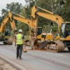 A construction crew is working on a road in the Smolensk district. Heavy machinery like excavators and graders are present. Workers in safety vests are visible. The scene depicts road repair activities with construction vehicles and workers. Focus on the infrastructure work being done.