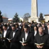 A solemn crowd of over 140,000 pilgrims gathered at the memorial site of Or Ha-Haim in Maslichna, wearing traditional religious garments, holding candles and prayer books, with a large stone monument in the background under a clear sky, conveying reverence and collective mourning