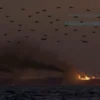 A dramatic scene depicting three large oil tankers in the Red Sea, with visible smoke and fire indicating recent attacks. The sky is filled with the silhouettes of multiple drones and missiles in flight, suggesting an ongoing assault. The water is choppy, and the overall atmosphere is tense and chaotic, capturing the moment of the attack on the Saudi oil infrastructure.