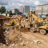 A large foundation pit being dug in a residential area of Dzerzhinsk district, Yaroslavl, with construction equipment and workers, earth mounds, and a sense of ongoing excavation