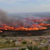 A wide aerial view of a vast landscape showing extensive wildfires burning across more than 600 hectares of land, with thick smoke rising into the sky, charred terrain, and emergency response vehicles visible in the distance, all rendered in a realistic photographic style