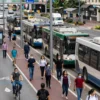 A bustling city scene with people commuting using various modes of transportation such as buses, trams, and bicycles, highlighting the growth in public transit usage. Include a diverse crowd of passengers, modern vehicles, and urban infrastructure like bus stops and bike lanes. Show a sense of movement and activity.