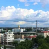 A cityscape of Izhevsk with a mix of summer warmth and rain. The image should depict a typical summer day in the city, with some areas showing sunshine and warmth, while others are under light rain. The scene should include buildings, streets, trees, and people going about their day, some with umbrellas. The sky should be partly cloudy with patches of blue, indicating the transition between rain and sunshine.