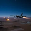 A military aircraft flying over a desert landscape at night, with explosions and smoke visible in the distance, under a starry sky. The scene should depict a covert military operation with no visible text or prohibited content.