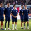 A group of French national football team players standing together on a grassy field, wearing blue jerseys with white and red accents, looking solemn and dignified, with the French flag subtly visible in the background, soft natural lighting, realistic facial expressions, no text or symbols on jerseys or background