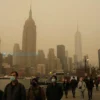 A panoramic view of New York City covered in a thick yellow smog due to wildfires in Canada. The skyline is partially obscured, with iconic buildings like the Empire State Building and One World Trade Center visible but hazy. The sky is a murky yellow, and the streets below are dimly lit with a yellowish tint. People are seen wearing masks and walking hurriedly, some looking up at the sky in concern.