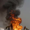 A massive oil field fire engulfing an Iraqi oil field, with towering flames and thick black smoke rising into the sky, burning oil derricks and storage tanks, intense orange and yellow fire contrasting with dark smoke, visible heat distortion, scattered debris, and a hazy atmosphere under a dark, smoky sky