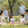 A happy family of six with two young children, a boy and a girl, playing in a sunny park. The parents are smiling and interacting with the kids, while the grandparents sit on a bench nearby, watching warmly. Include trees, a playground, and a picnic blanket with snacks. Show a joyful, lively atmosphere with natural lighting.