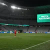 A football match between Mexico and England in the round of 16 of the World Cup, with an empty stadium under dim lighting, a large digital scoreboard showing 'MATCH POSTPONED' in bold, and rain falling gently on the pitch