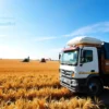 A wide-angle view of golden wheat fields in the Volgograd region under a bright blue sky, with modern combine harvesters working in the distance and a large cargo truck loaded with grain bags in the foreground, symbolizing agricultural export growth